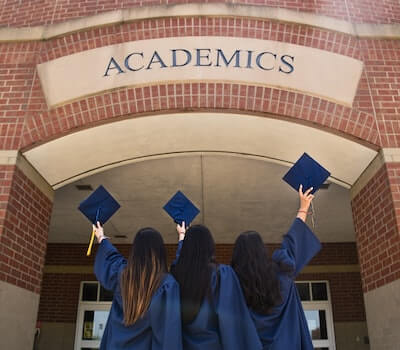 Students holding graduation caps in front of a school