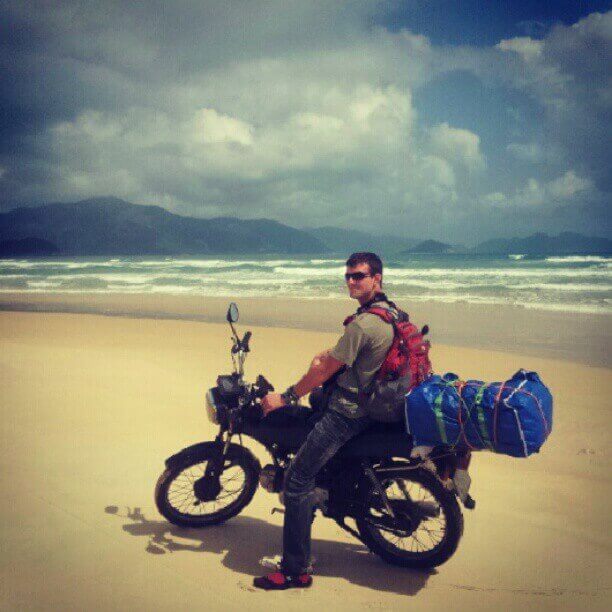 man sitting on motorbike with backpack on beach